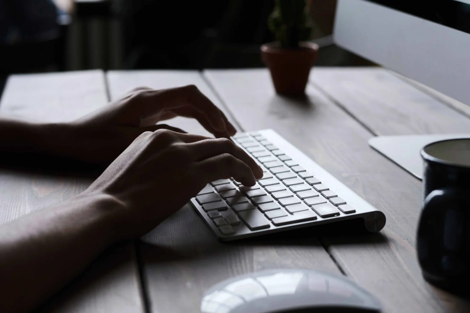 Person typing on a wireless keyboard at a wooden desk beside a monitor.