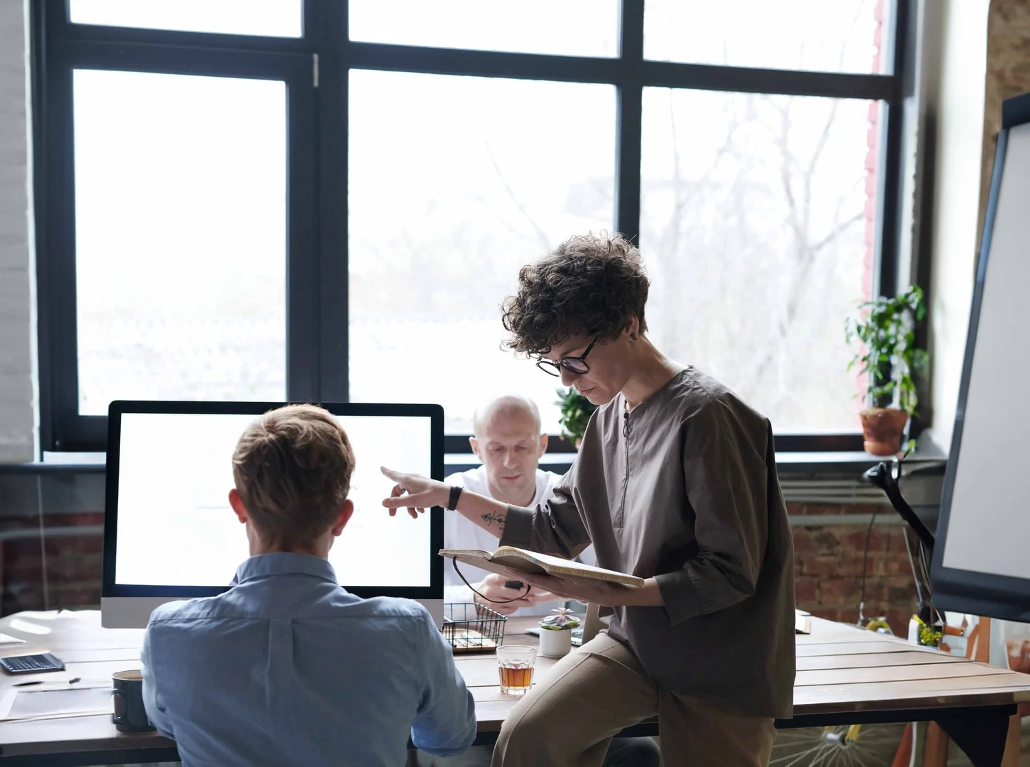 A woman standing and pointing at a man seated at a desk in an office with large windows, two computer monitors, and plants.