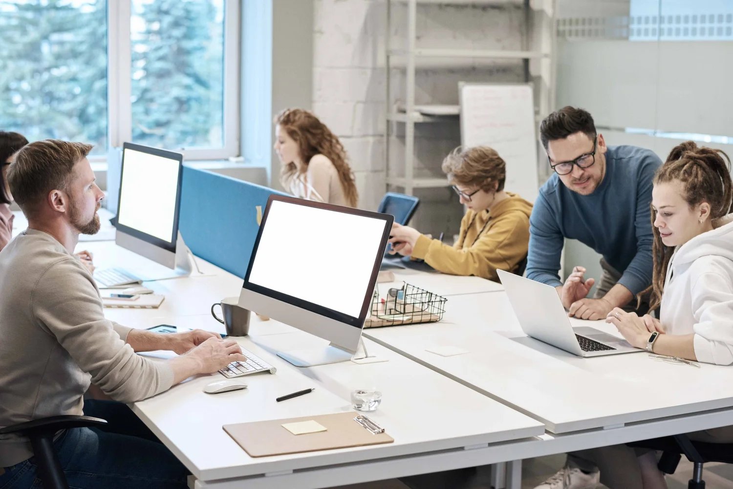 Group of young people working in a modern office space with computers and laptops at a shared desk.
