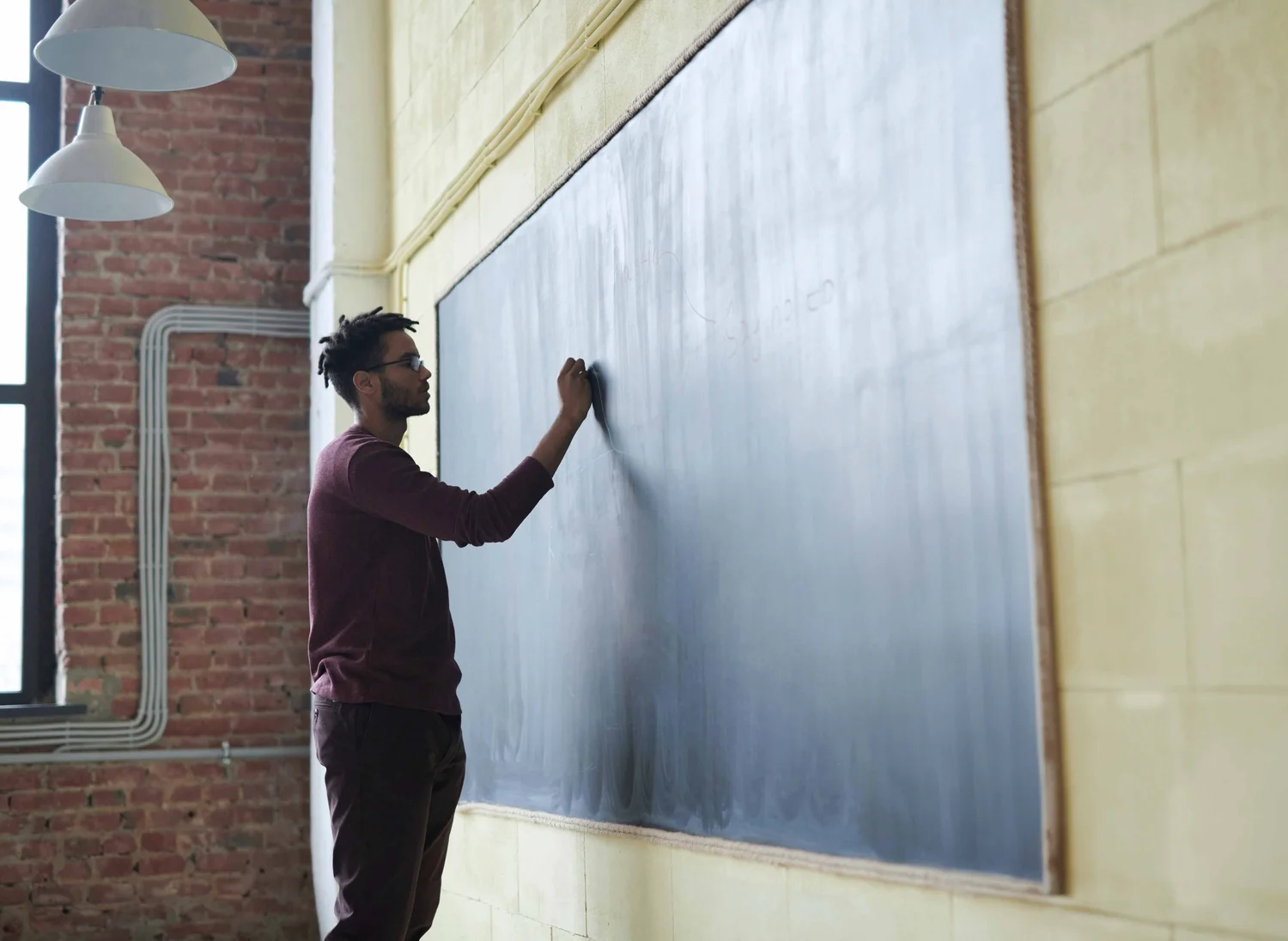 A man with glasses and a maroon sweater writing on a large chalkboard in a classroom with exposed brick wall and yellow paneling.