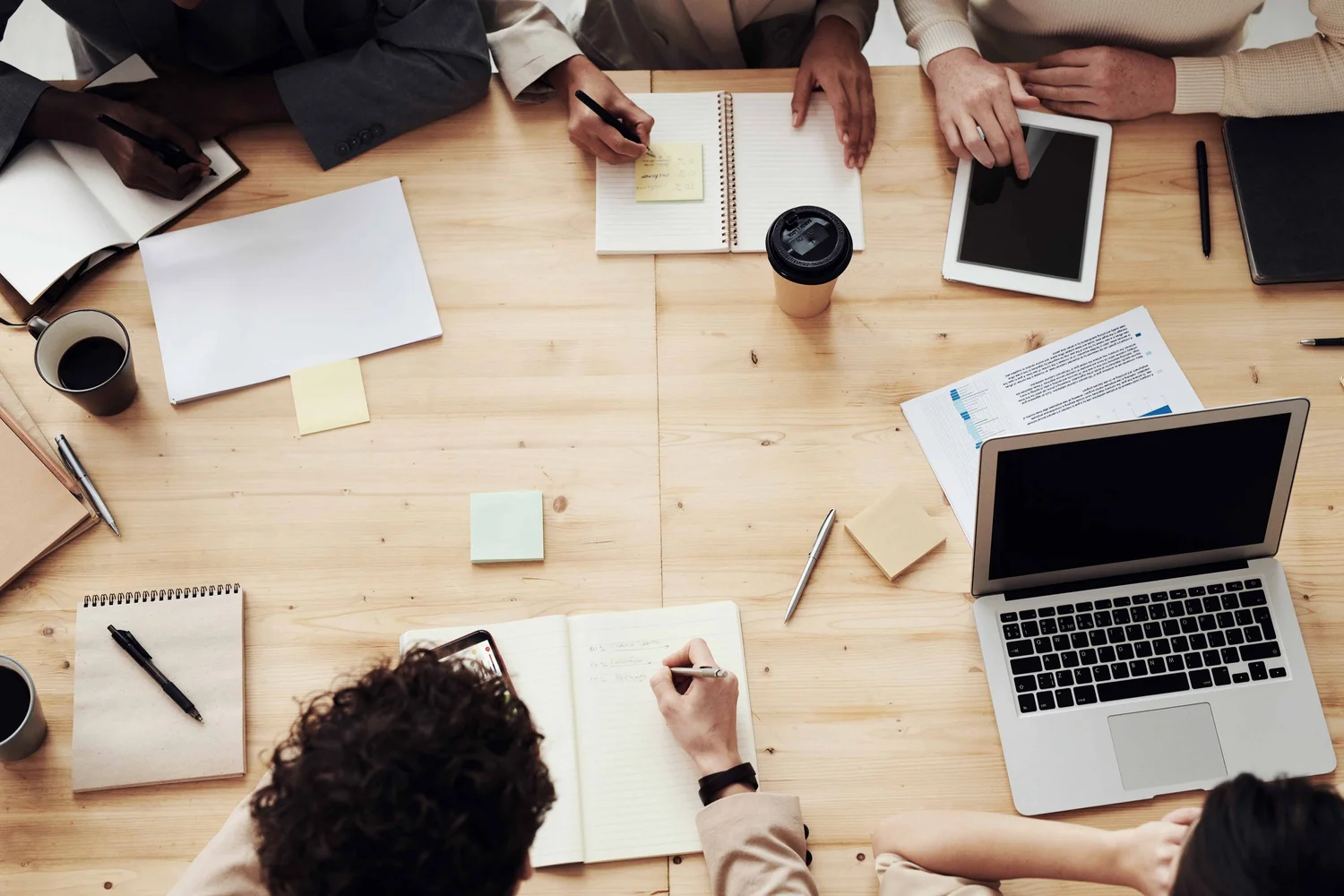 Top-down view of a group of people working at a wooden table with laptops, notebooks, papers, pens, sticky notes, and cups of coffee.