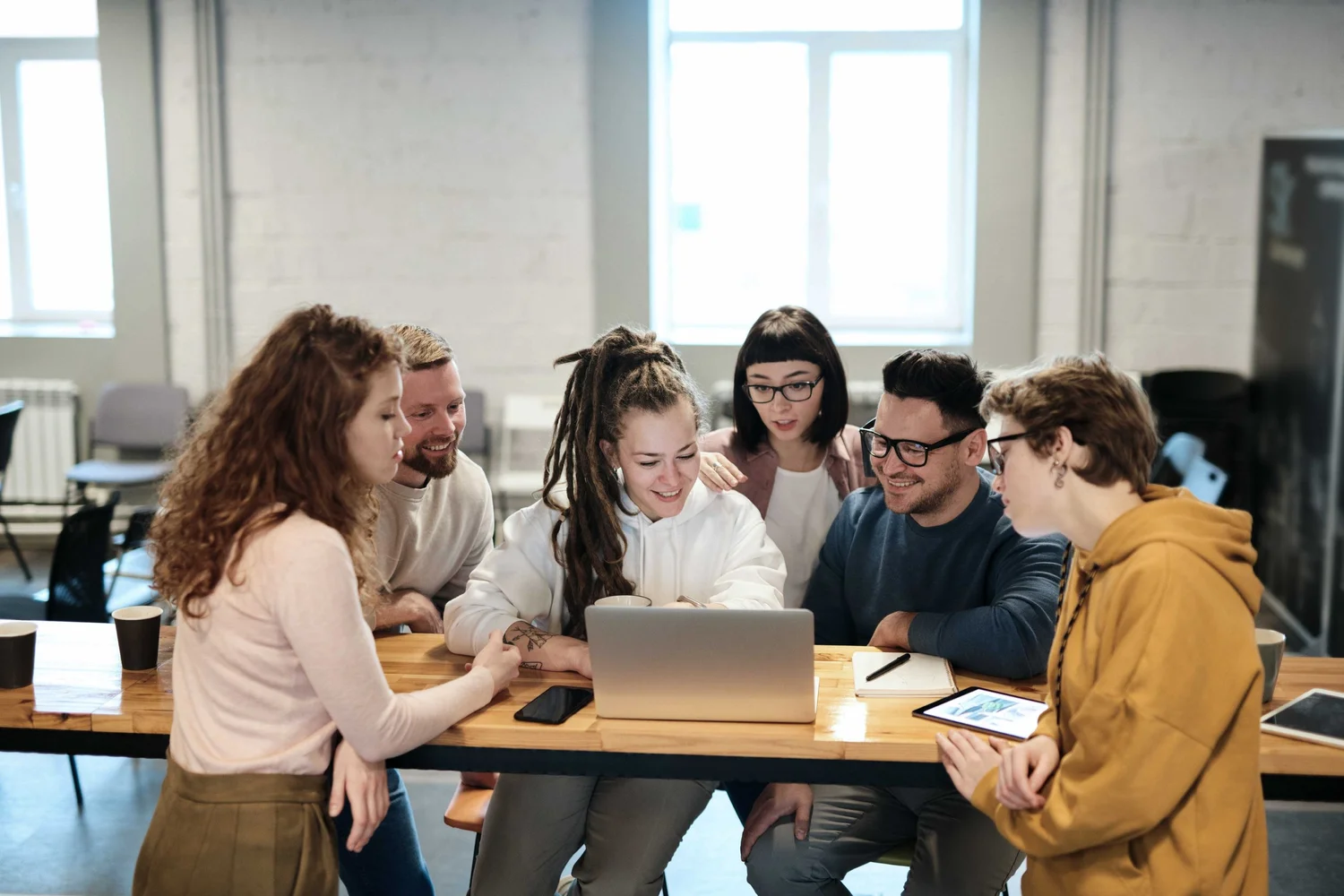 Group of six diverse young adults gathered around a table, smiling and looking at a laptop in a modern, well-lit office.